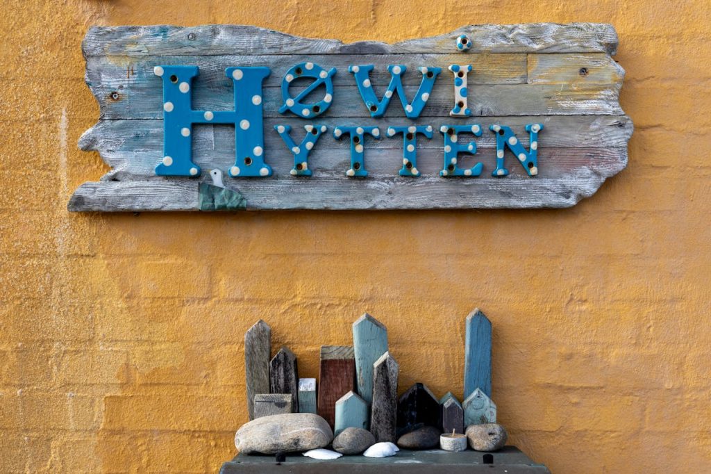 A rustic wooden sign reading 'Høwi Hytten' mounted on a yellow textured wall accompanied by small crafted wooden houses.