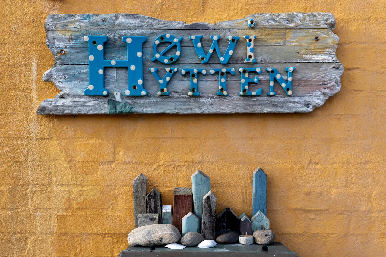 A rustic wooden sign reading 'Høwi Hytten' mounted on a yellow textured wall accompanied by small crafted wooden houses.